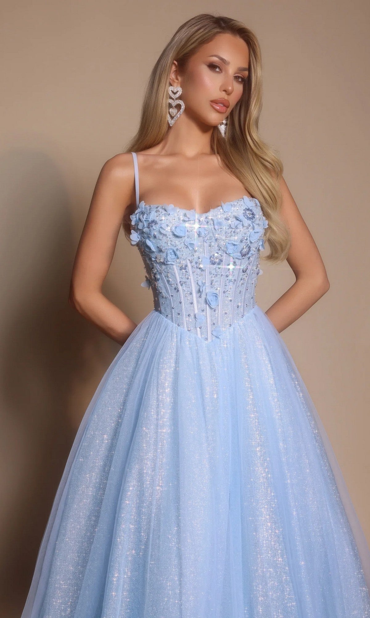 A woman in the Portia and Scarlett PS26146 Glitter Prom Ball Gown, a light blue tulle dress with floral accents on the boned-corset bodice and a shimmery skirt, stands against a plain beige background.